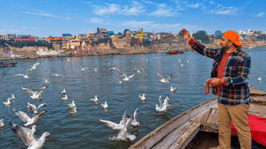 Dr. Vijay Kumar standing on a traditional wooden boat during sunrise on the River Ganga in Varanasi, surrounded by white birds with the historic ghats and ancient temples in the background.